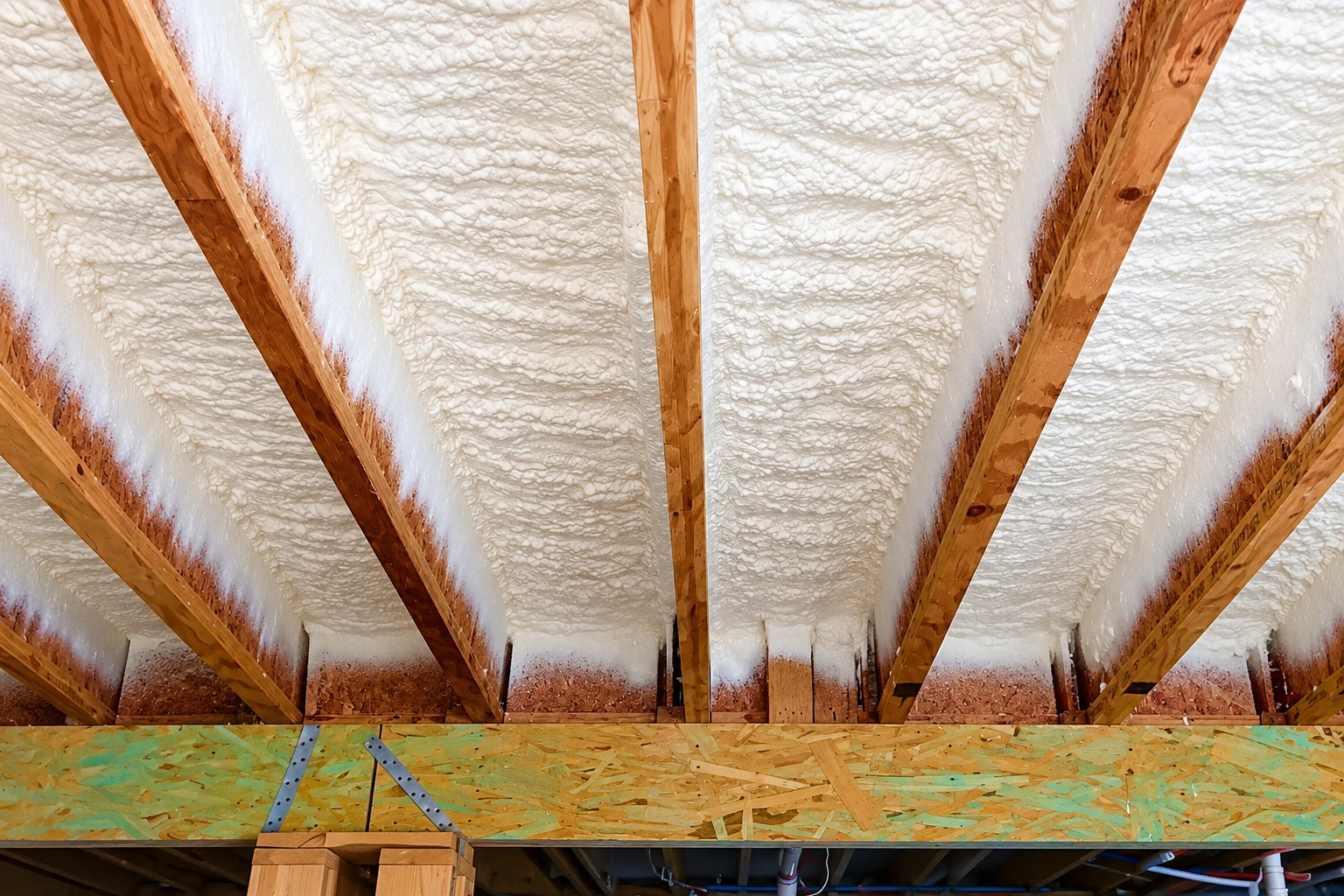 Spray foam insulation fills the spaces between wooden attic rafters above a plywood floor.