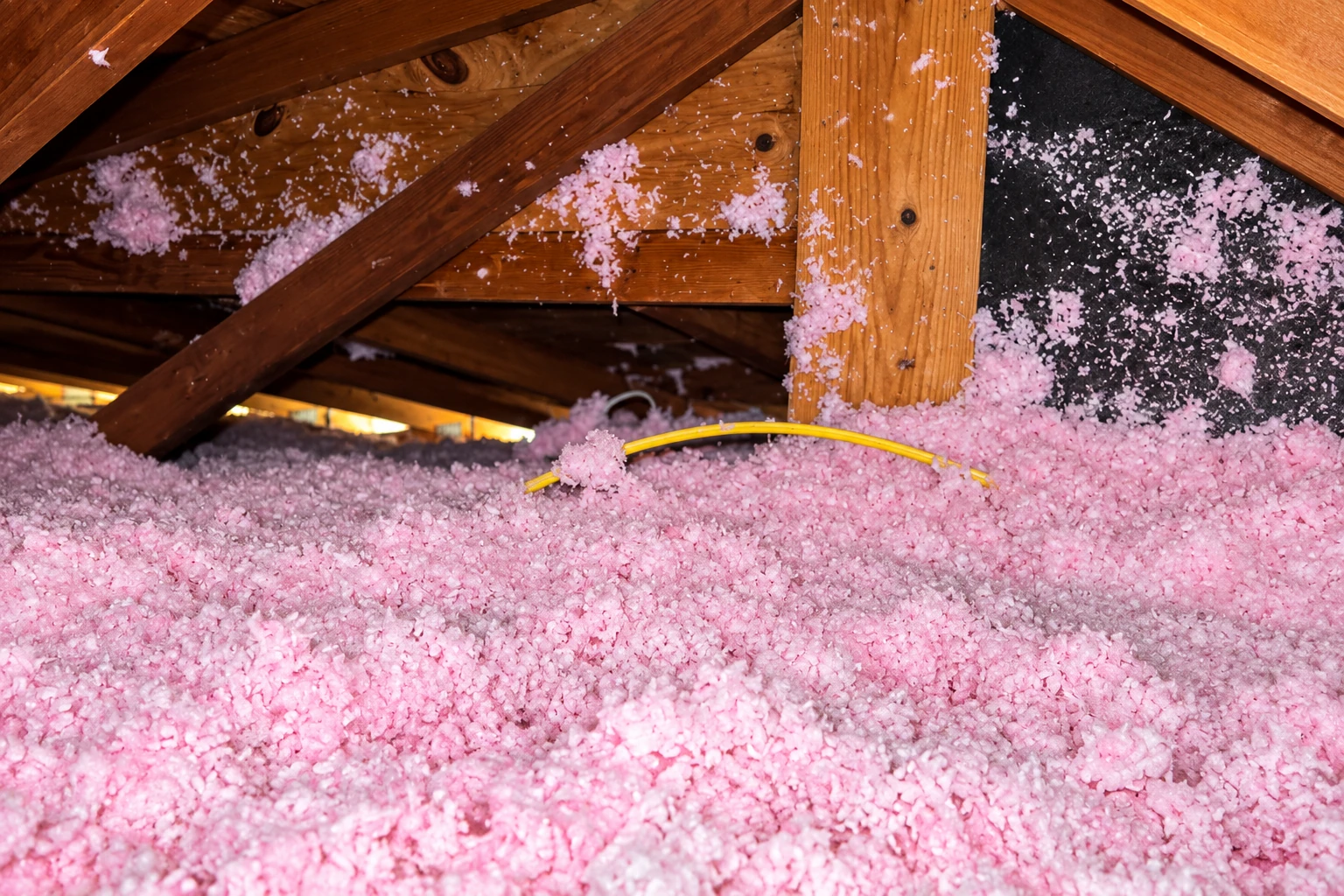 Pink blown-in insulation covers an attic floor around exposed rafters and a yellow cable.