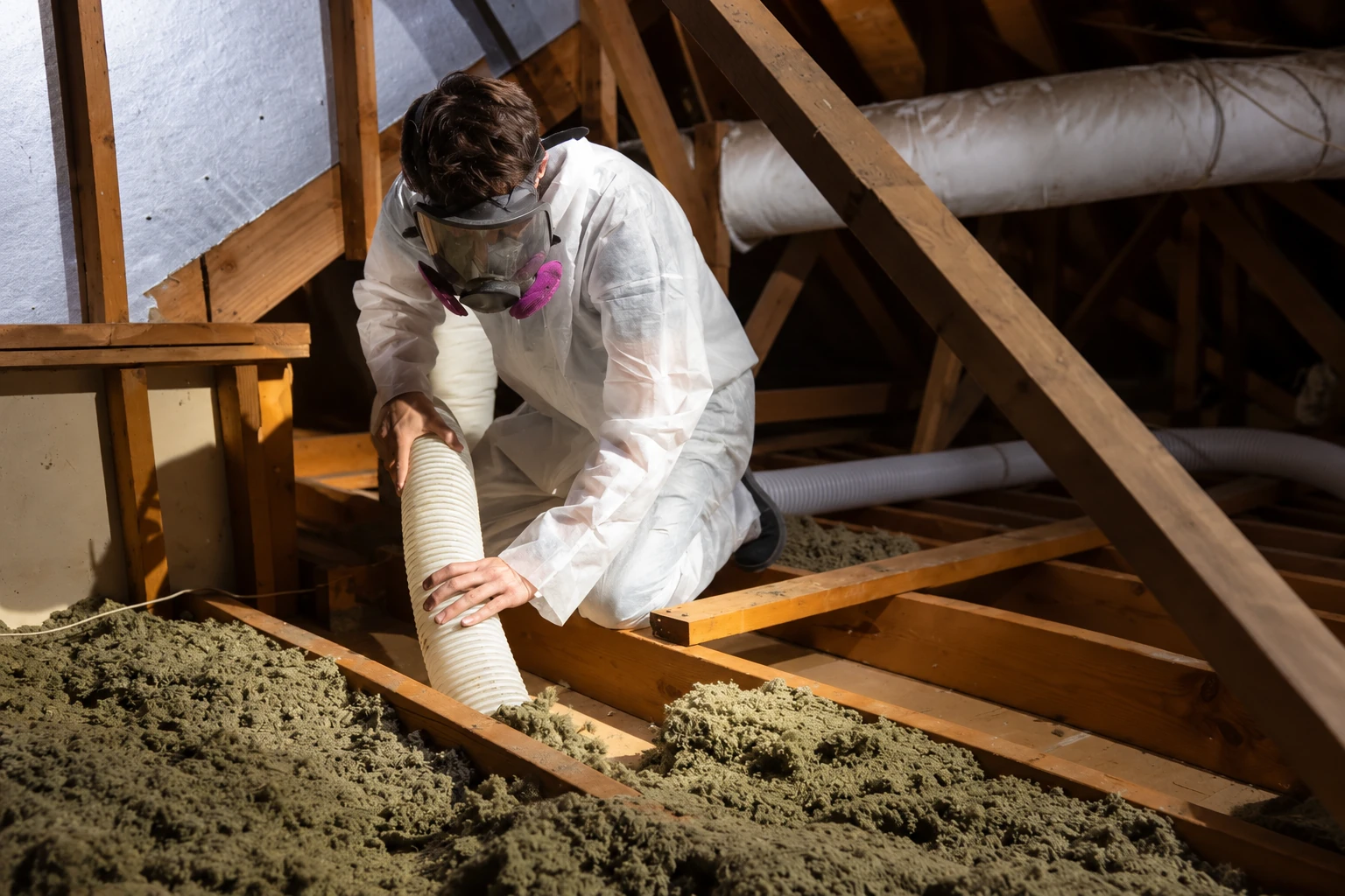 A worker in protective coveralls and a respirator removes loose attic insulation using a flexible hose.